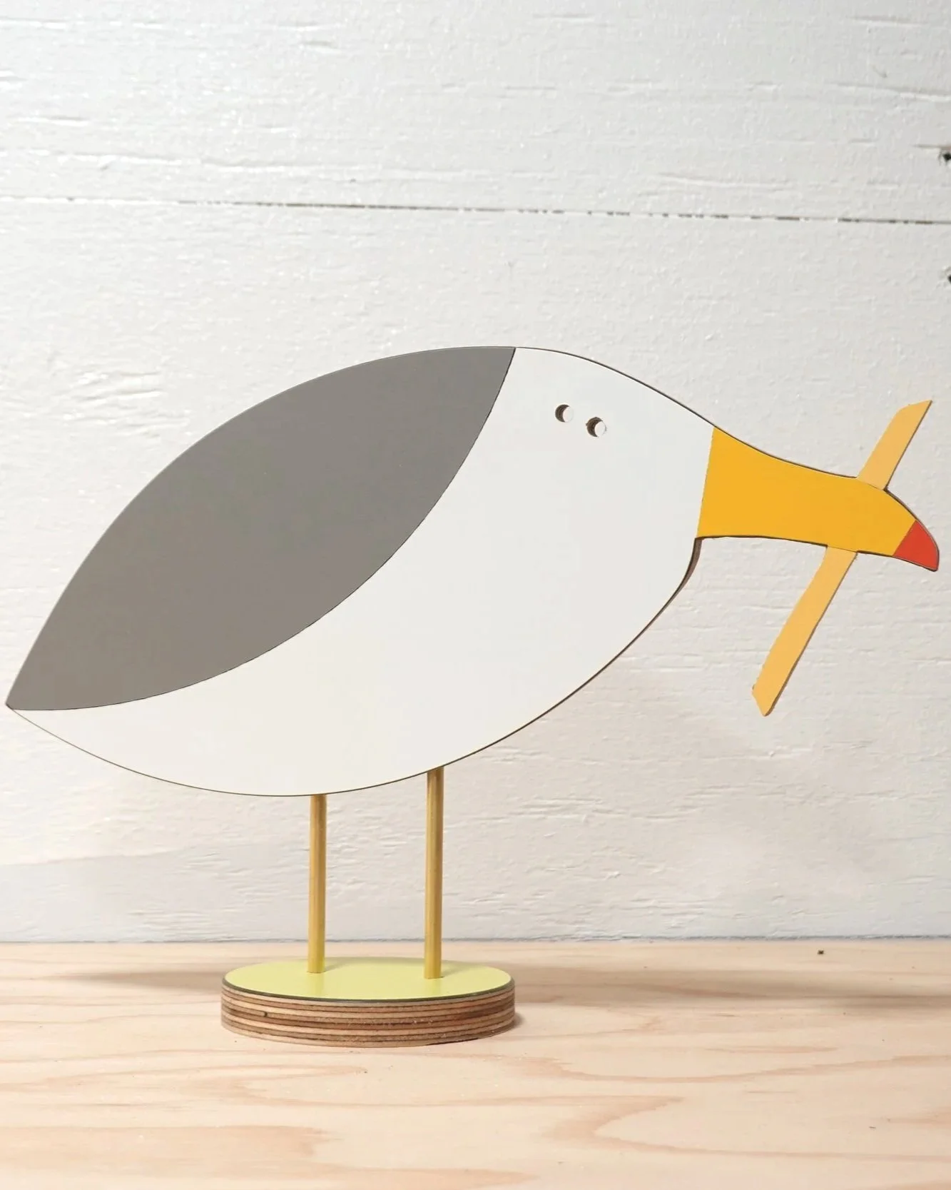 Wooden bird figure with a yellow beak and gray and white body, standing on a circular wooden base.