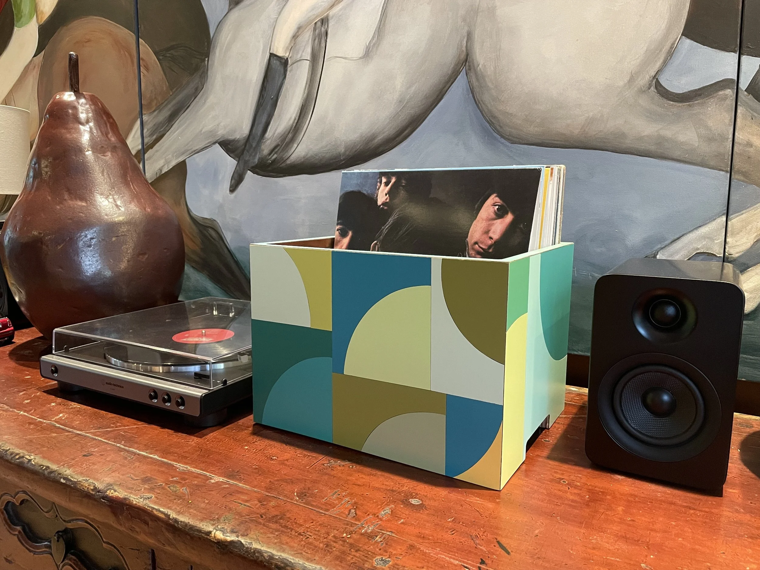 A tabletop with a turntable, a colorful record storage box with records inside, and a black speaker, with a large painted horse mural in the background.
