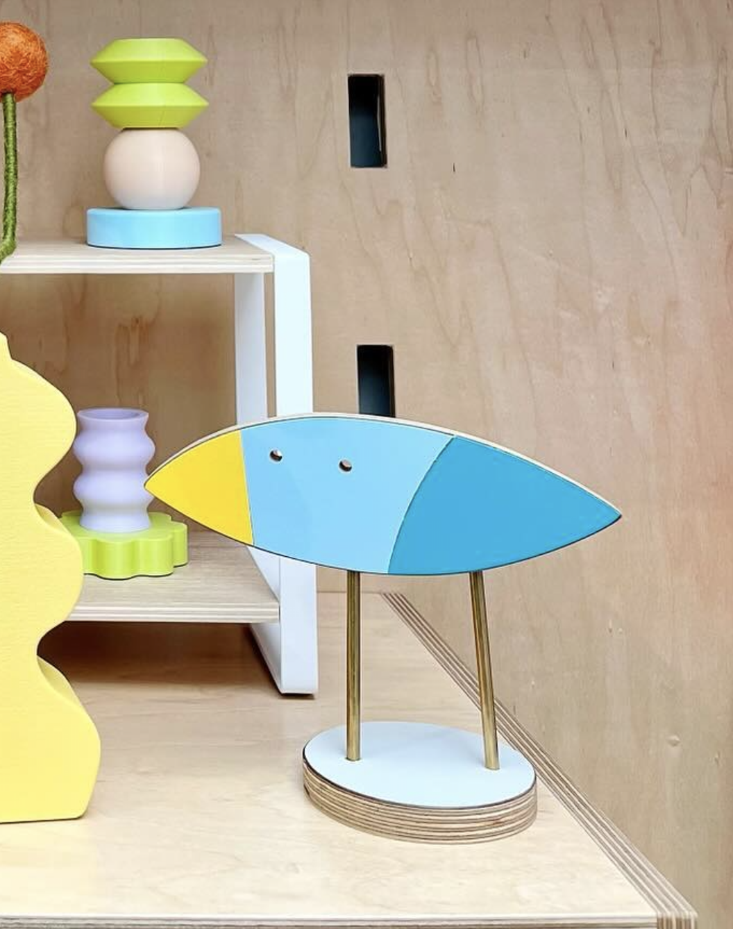 Colorful decorative fish-shaped wooden sculpture with blue, yellow, and light blue colors, sitting on a white and wood base with gold legs, in a room with wooden walls and assembled furniture.