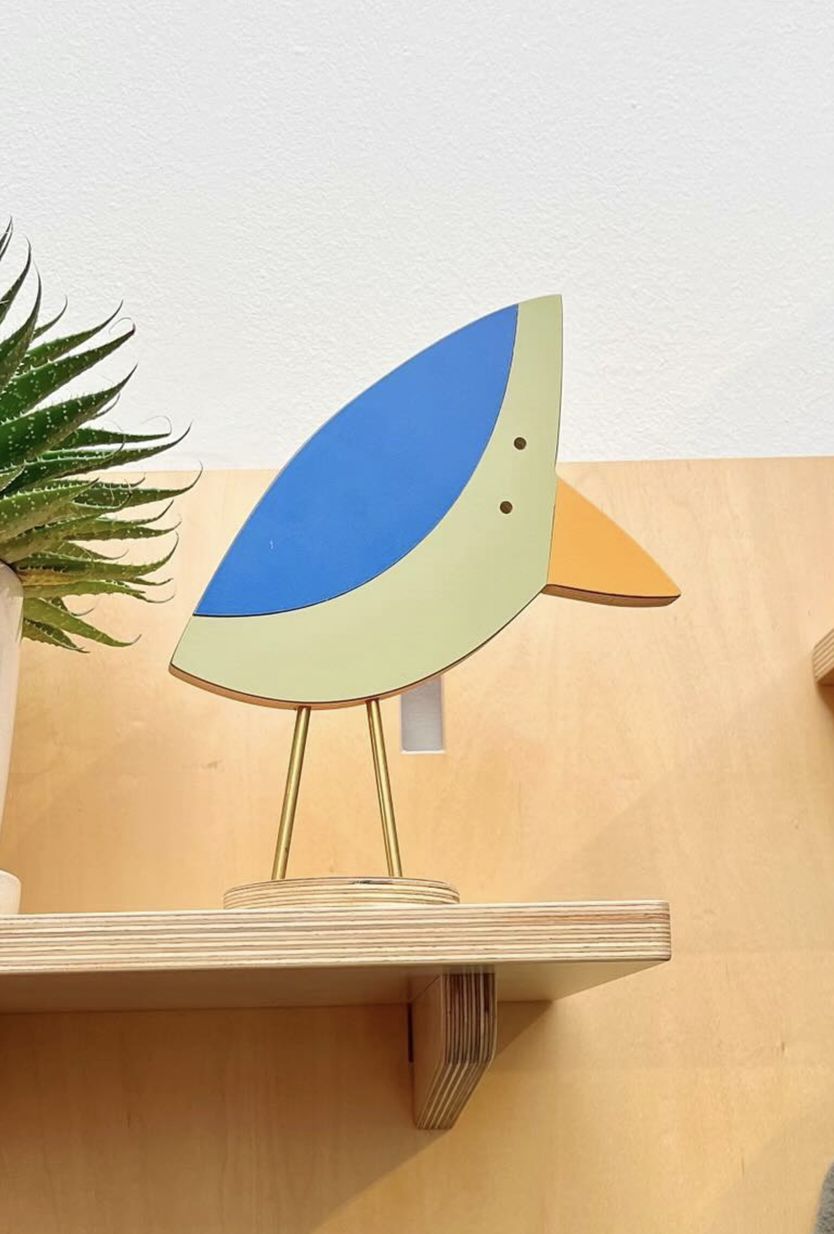 Decorative bird sculpture with a green body, blue wing, and orange beak on a wooden shelf with a plant nearby.