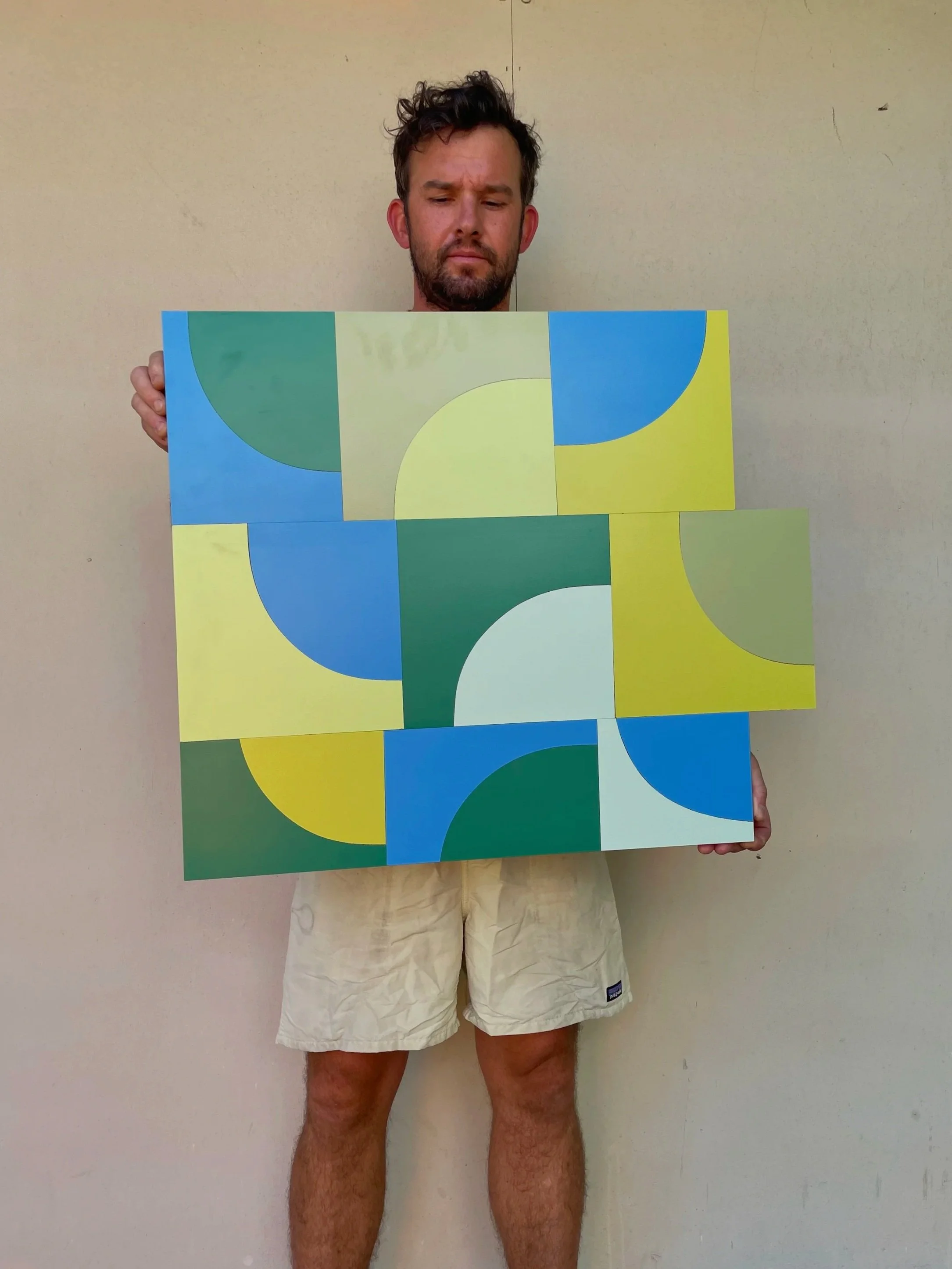 A man holding a colorful abstract geometric painting with various shapes and colors including green, blue, yellow, and white, standing against a plain wall.