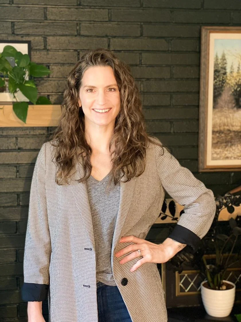 Woman with curly brown hair smiling, wearing a beige and black checked blazer over a grey shirt, standing indoors against a black brick wall with framed artwork and a plant in a white pot in the background.