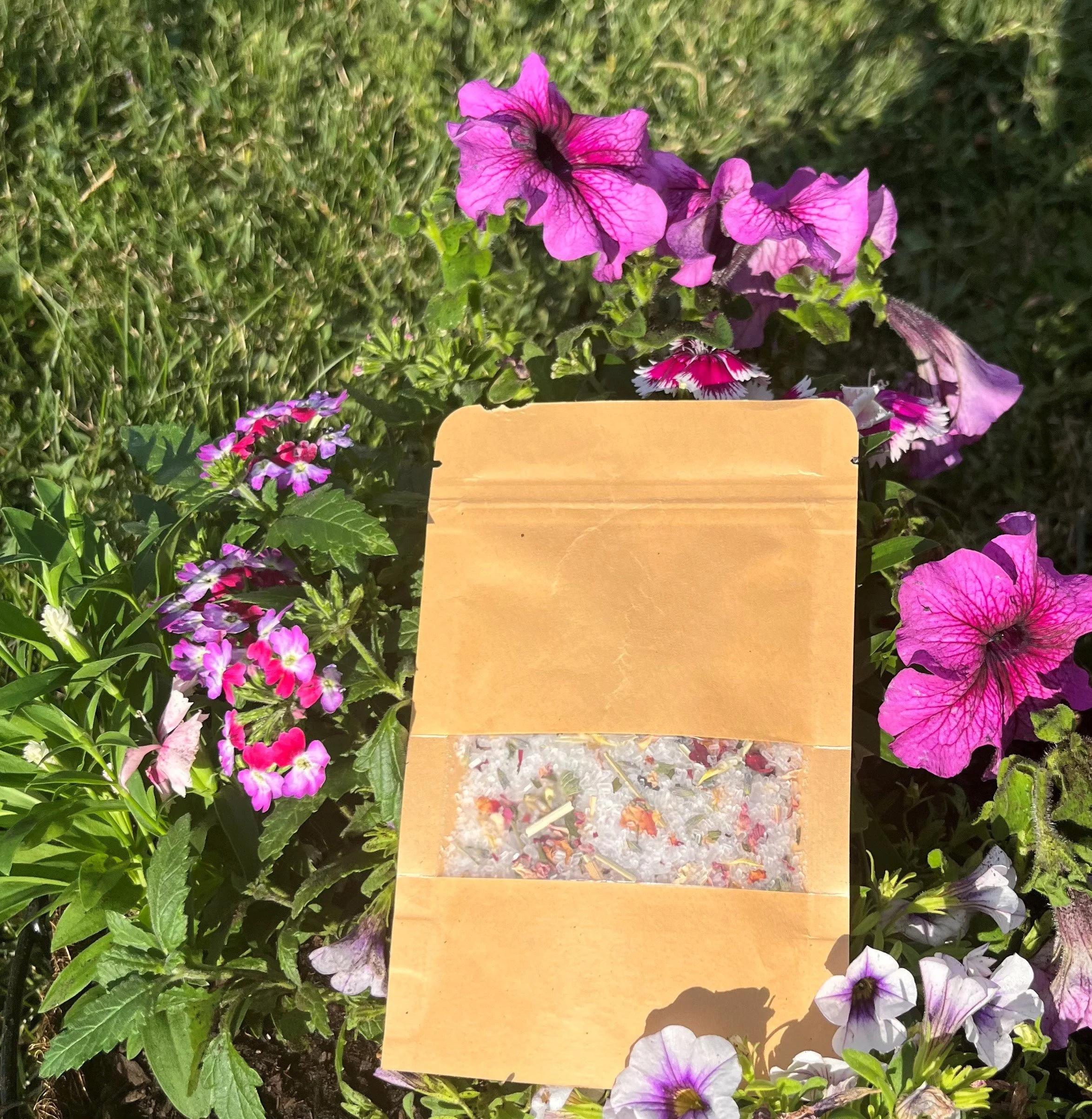 Colorful flowers in pink, purple, and white surrounding a small paper bag filled with what appears to be crushed ice and flower petals, set in a garden.