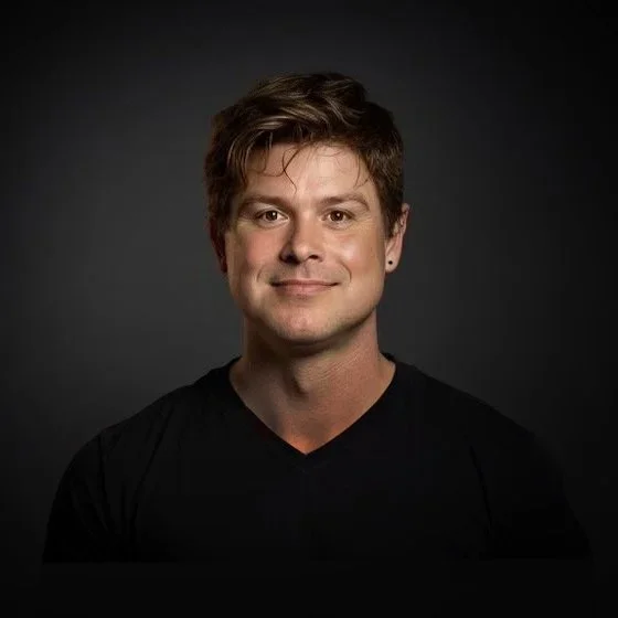 Portrait of a young man with short brown hair, wearing a black shirt, smiling, against a dark gray background.