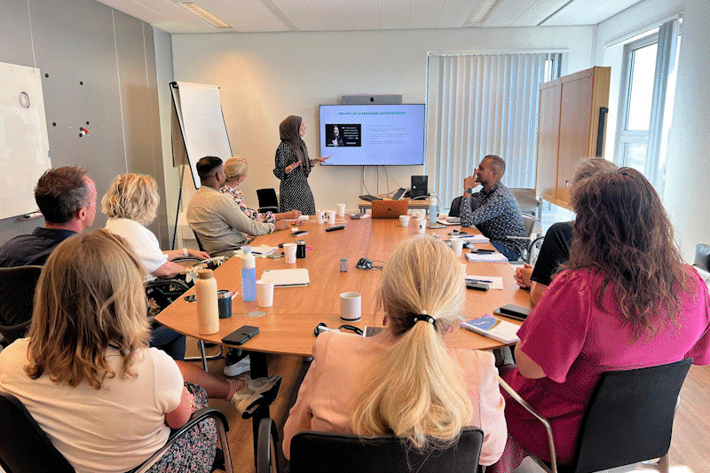 A group of diverse people attending a business meeting or presentation in a conference room, with a woman standing at a screen at the front.