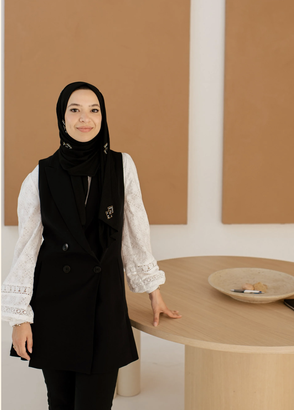 A woman wearing a black hijab and a black sleeveless coat with lace sleeves, standing indoors next to a round wooden table with a decorative bowl and some items on it.