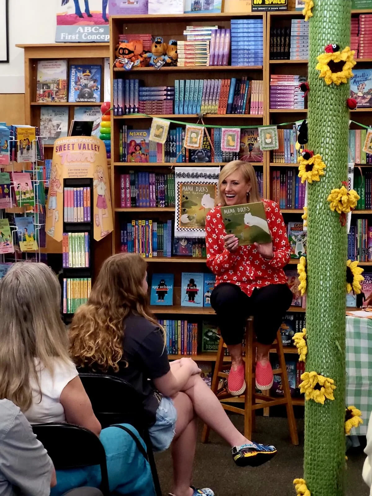 A woman sitting on a stool in a bookstore or library, reading a children's book titled "Mama Duck" to a group of children. The shelves behind her are filled with colorful books, and there is a decorative green post with yellow flowers and plush ladybug decorations on the right side of the image.