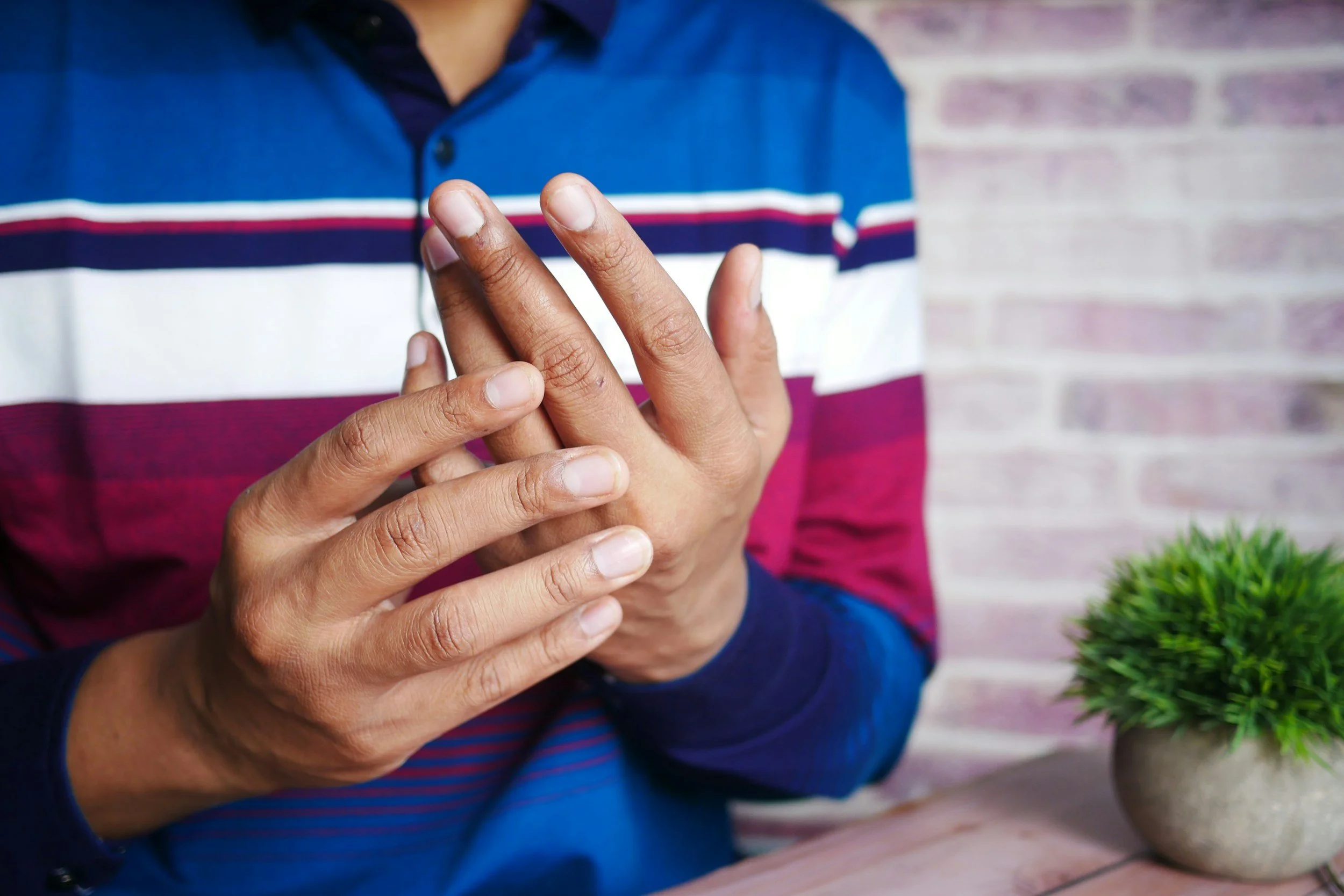 A person rubbing their hands together, wearing a long sleeve shirt with blue, white, red, and purple stripes. A small green potted plant is on a table beside them, with a brick wall in the background.