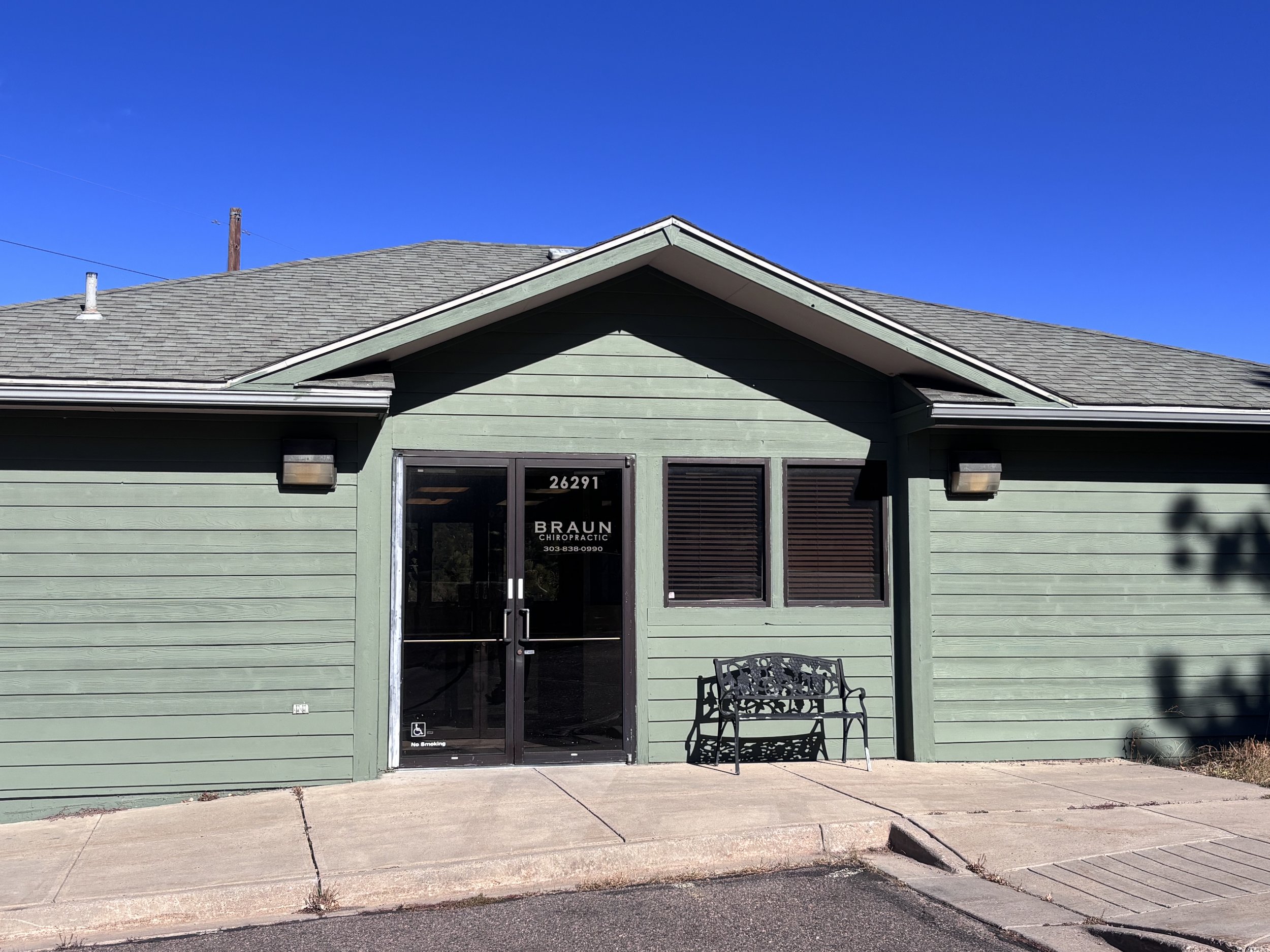 Green building with a glass door and two windows, promoting Braun Chiropractic. Bench outside on sidewalk under clear blue sky.