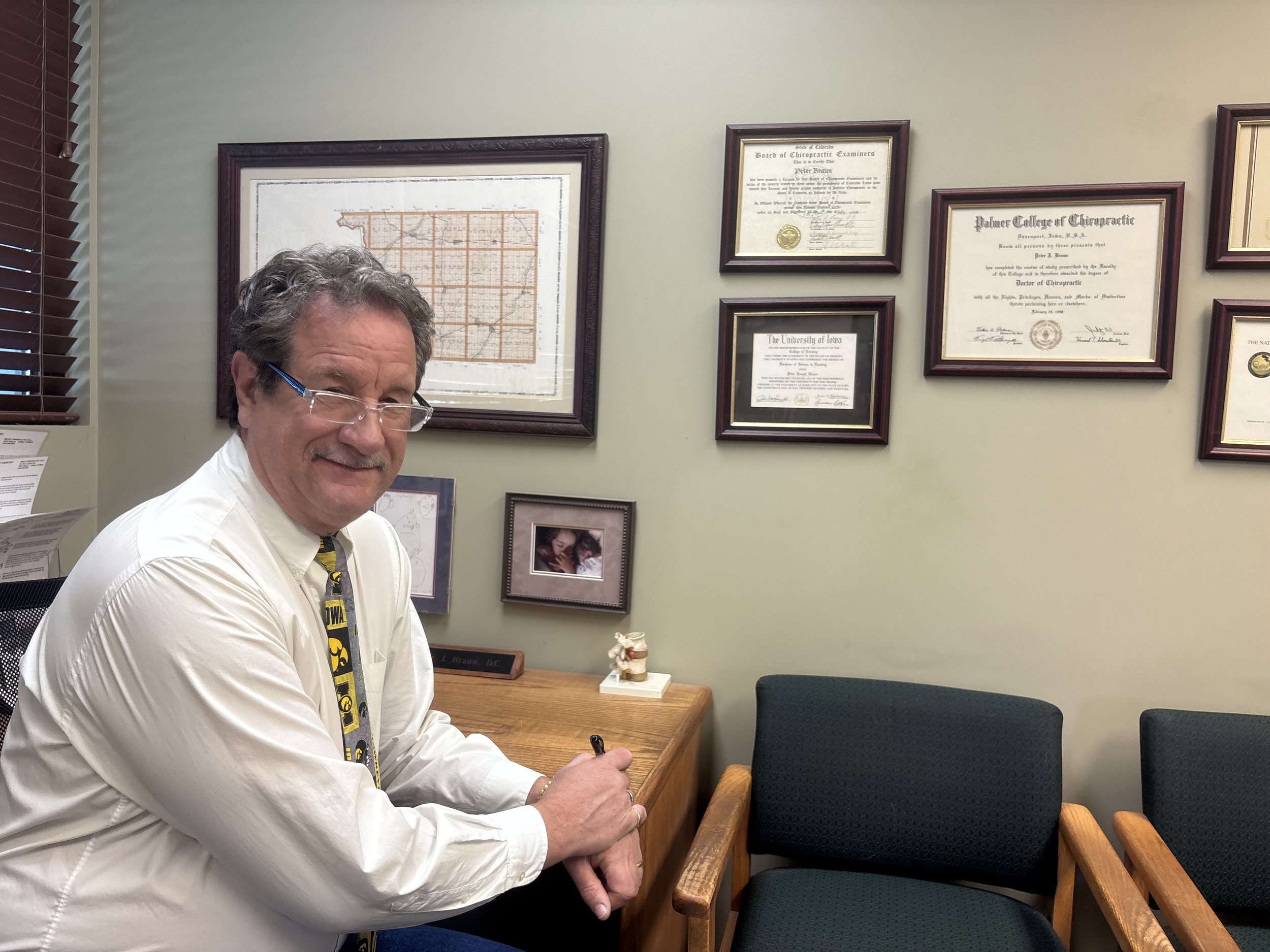 A smiling middle-aged man with glasses, wearing a white shirt and a black and yellow ties, is sitting at a wooden desk in an office. The wall behind him has framed diplomas and certificates and a framed map. There are two green chairs with wooden arm