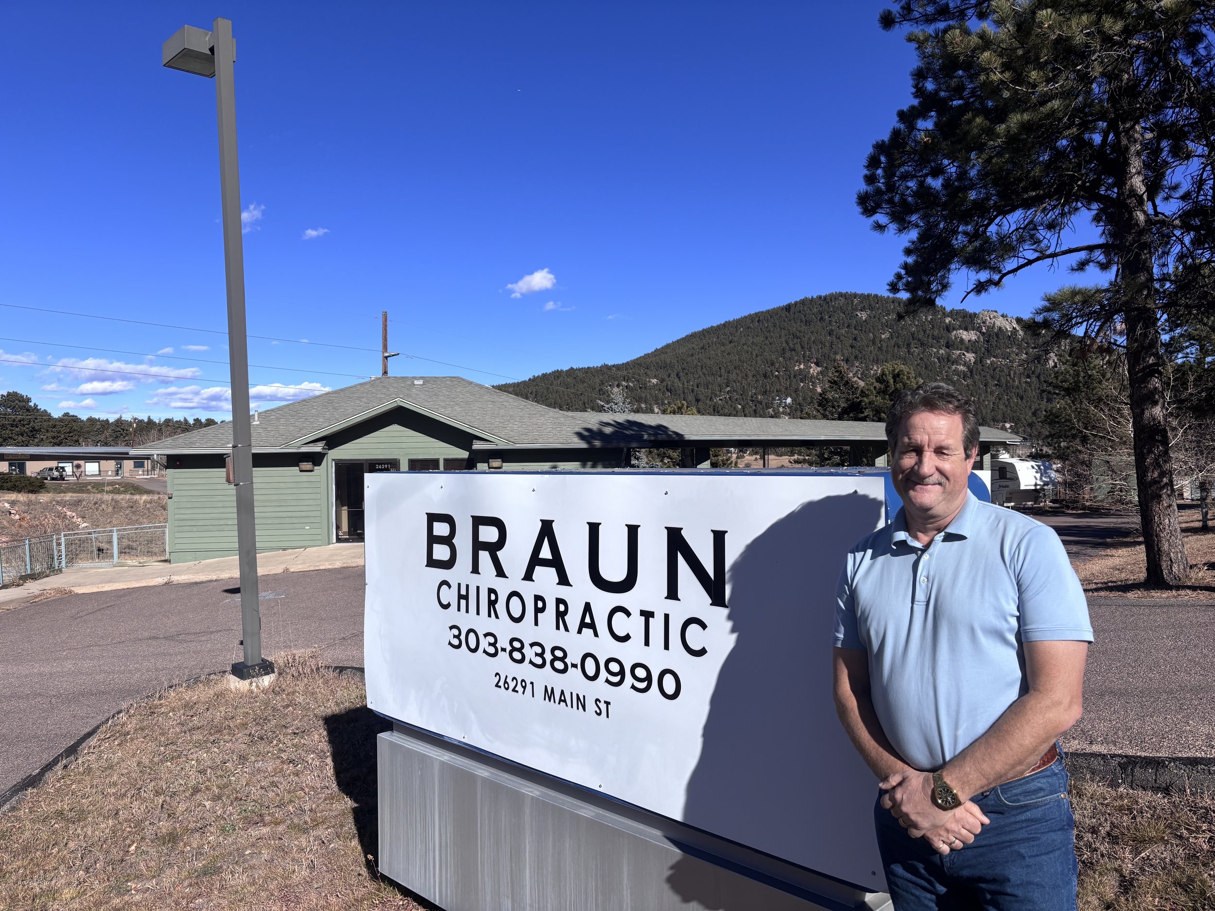 Man standing outdoors next to a large sign for Braun Chiropractic with a mountain in the background and a clear blue sky.