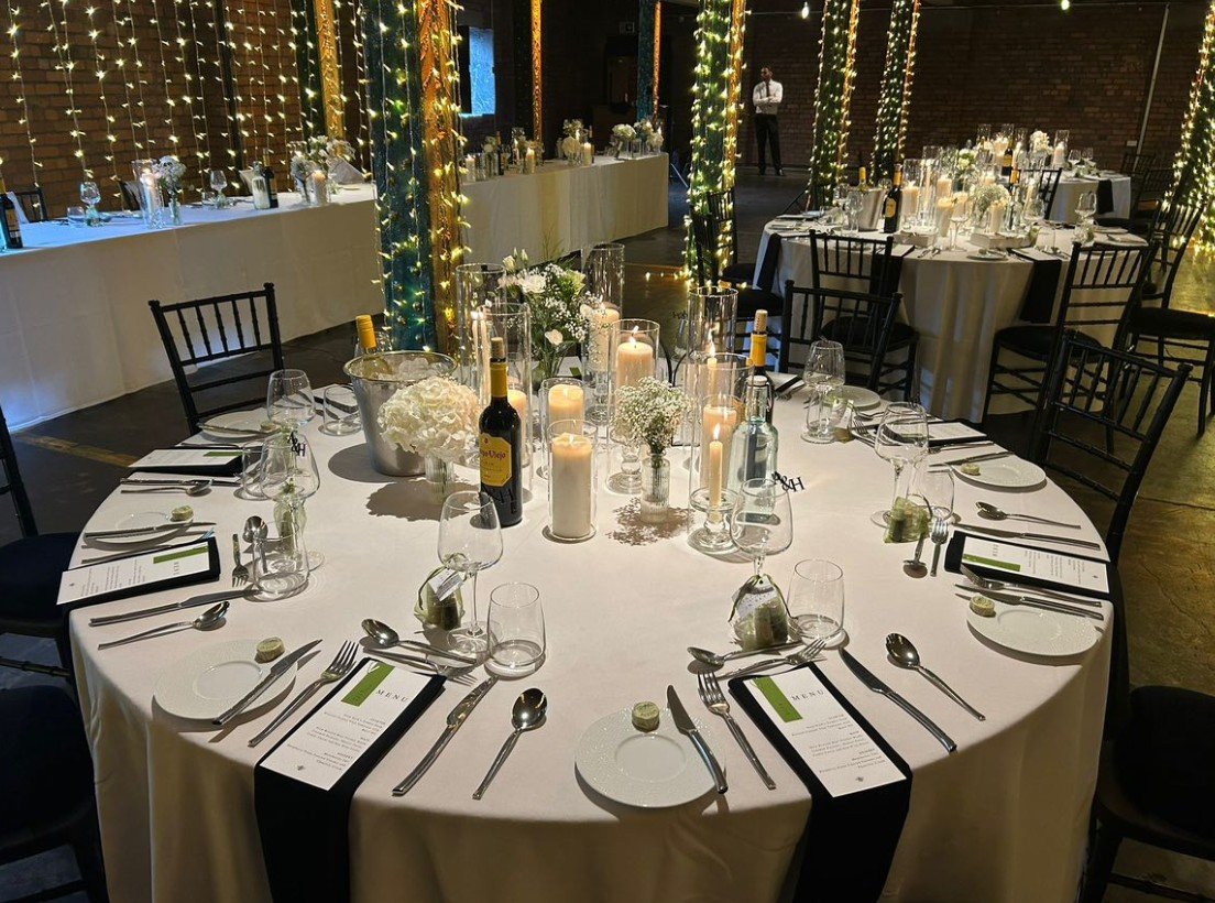 Wedding or event reception with round tables decorated with white tablecloths, candles, flowers, wine bottles, and place settings, with string lights and illuminated pillars in the background.