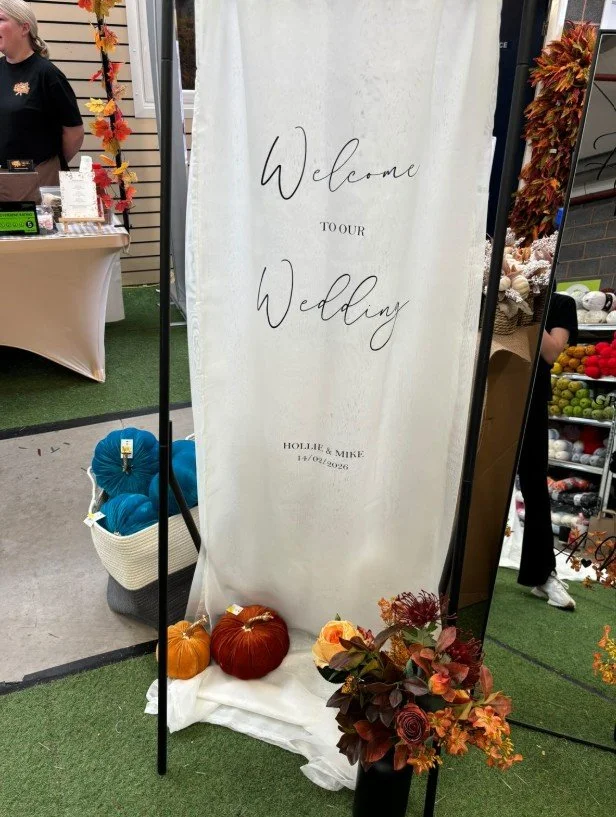 A white welcome sign at a wedding, reading 'Welcome to our wedding, Hollie & Mike, 14/01/2026,' surrounded by fall decorations including pumpkins, flowers, and foliage.