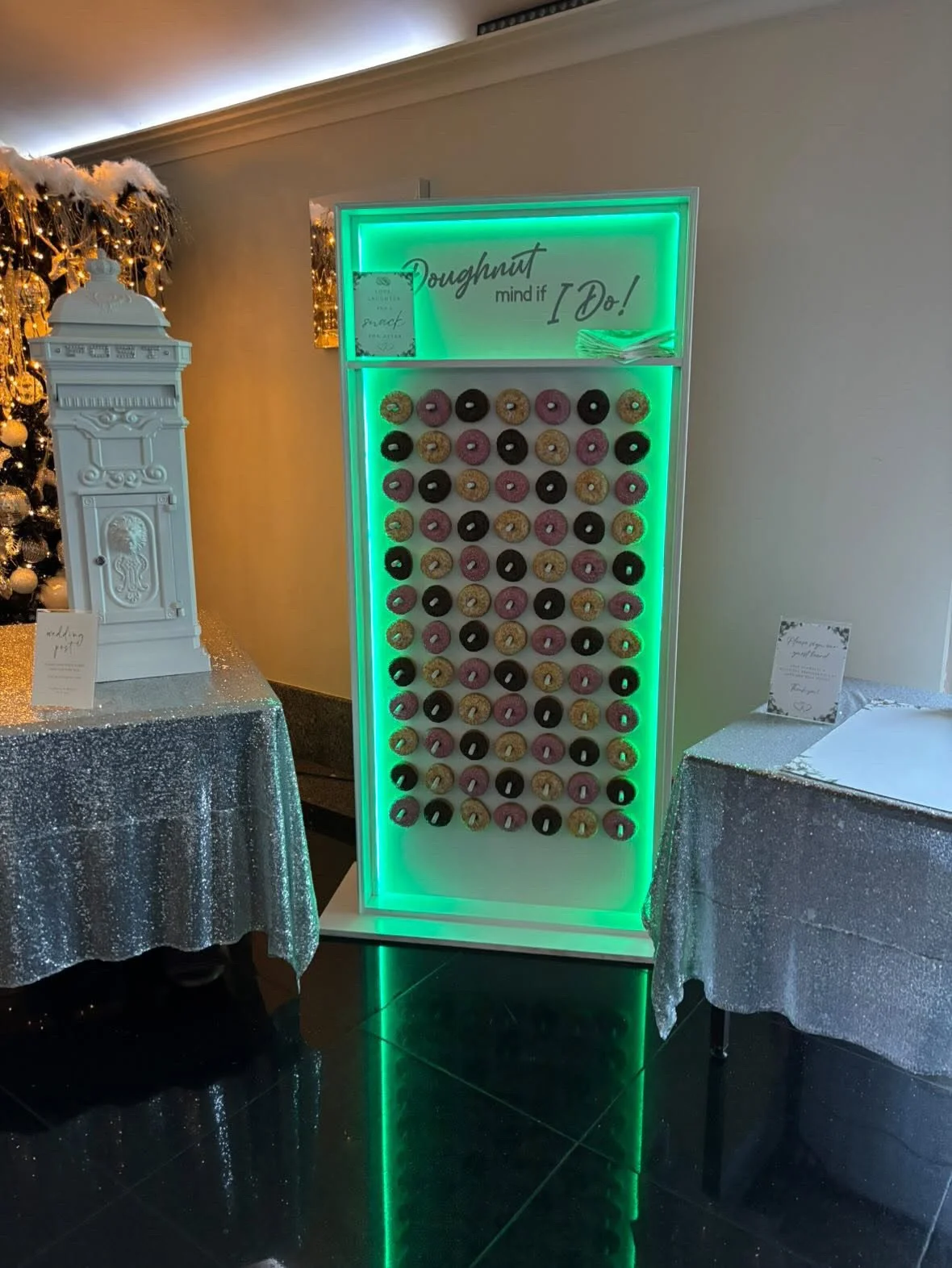 A donut wall with an arrangement of colorful donuts, decorated with a sign that reads 'Doughnut mind if I do!' in a neon-lit frame, situated between tables with sparkly tablecloths, likely at a festive event.