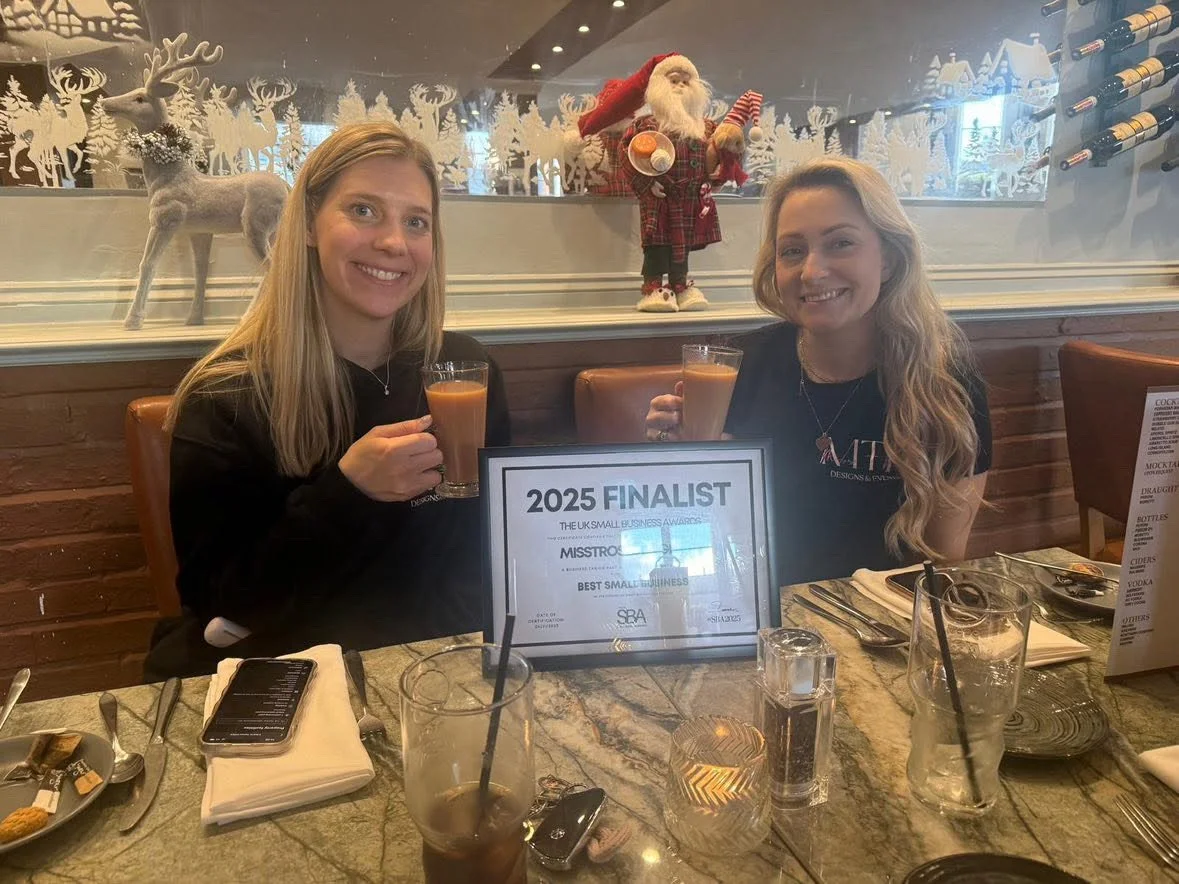 Two women smiling and holding drinks at a dining table with a framed award that reads "2025 Finalist, The UK Small Business Awards, Best Small Business." The background features Christmas decorations including a Santa Claus figure and reindeer.