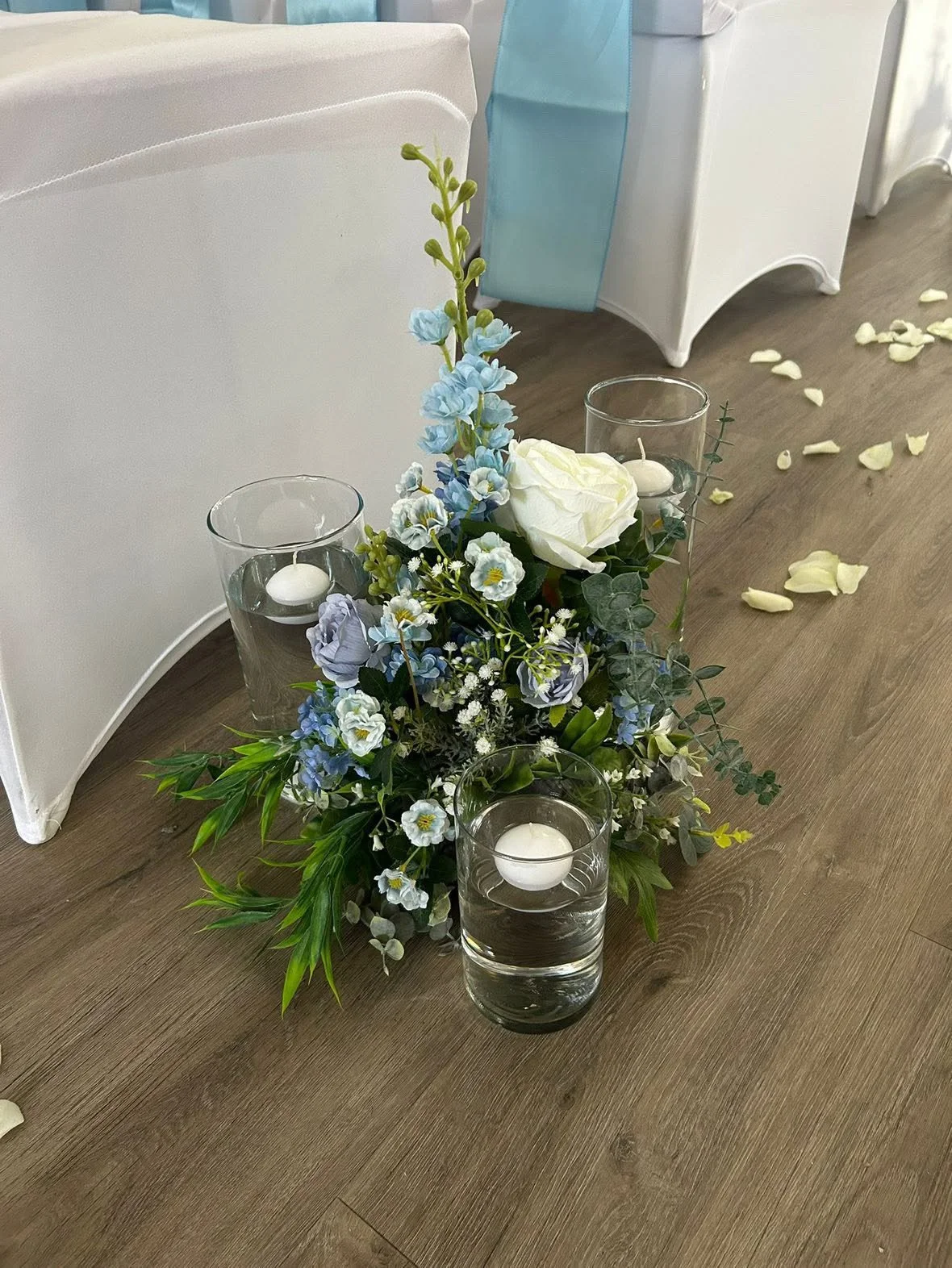 Floral centerpiece with white and blue flowers, green foliage, and three glass candle holders with floating candles on a wooden table surrounded by white chairs and scattered flower petals.