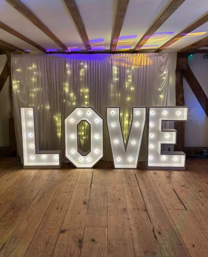 Large illuminated letters spelling out 'LOVE' against a backdrop of white curtains and yellow fairy lights, in a decorated indoor setting with a wooden floor and exposed beams.