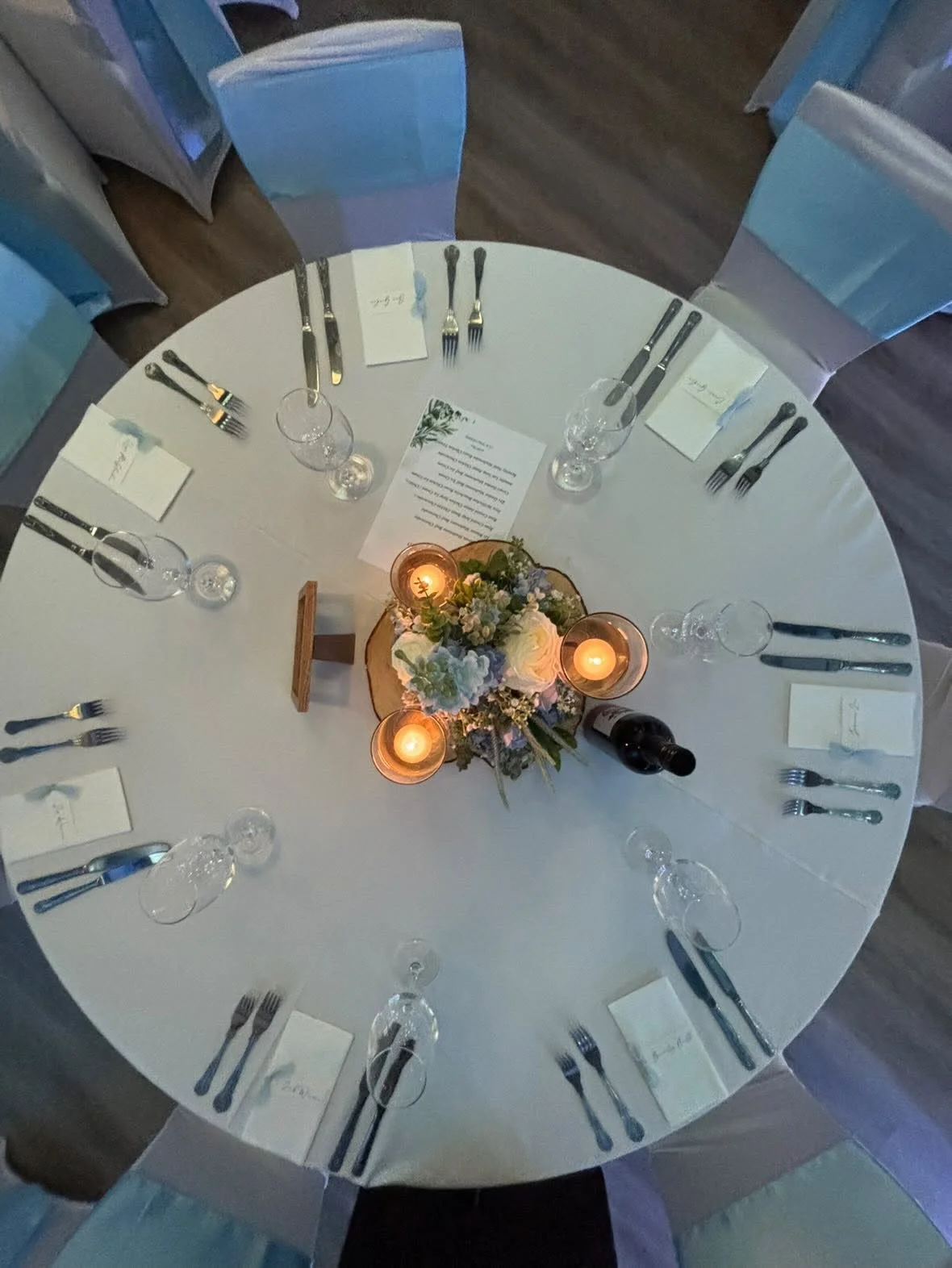Top-down view of a round table set for a formal dinner with a white tablecloth, floral centerpiece, candles, wine glasses, water glasses, and arranged silverware, with chairs covered in fabric.