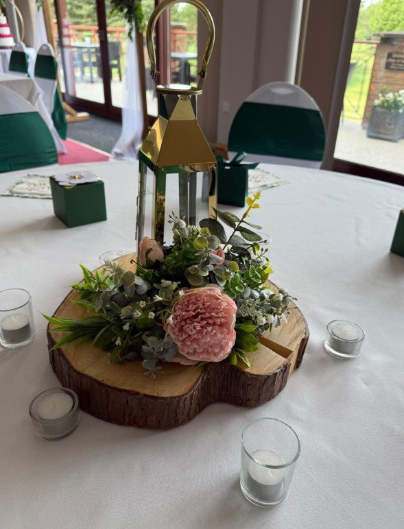 Decorative centerpiece on a white tablecloth, featuring a wooden slab, a gold lantern with a handle, and pink and green floral arrangements. Small white candles in glass holders surround the centerpiece, with a background of a room with large windows