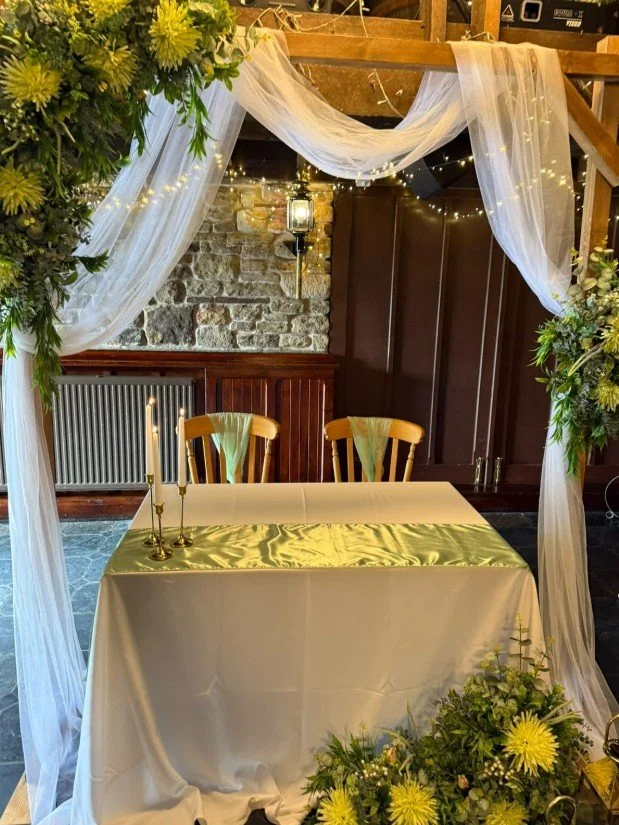 A decorated ceremony table with a white tablecloth and gold runner, surrounded by an arch of white tulle, greenery, and yellow flowers, set in a rustic indoor space with stone walls and fairy lights.