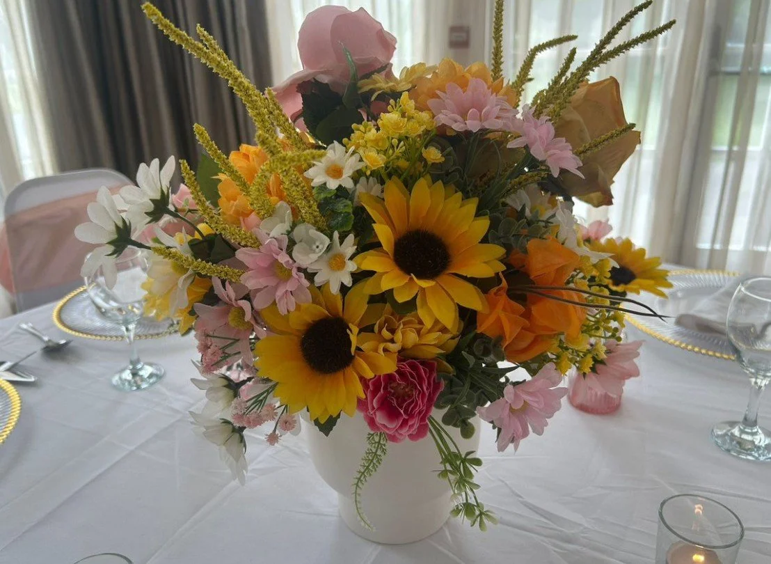 A flower centerpiece with sunflowers, pink roses, daisies, and other colorful flowers on a dining table.