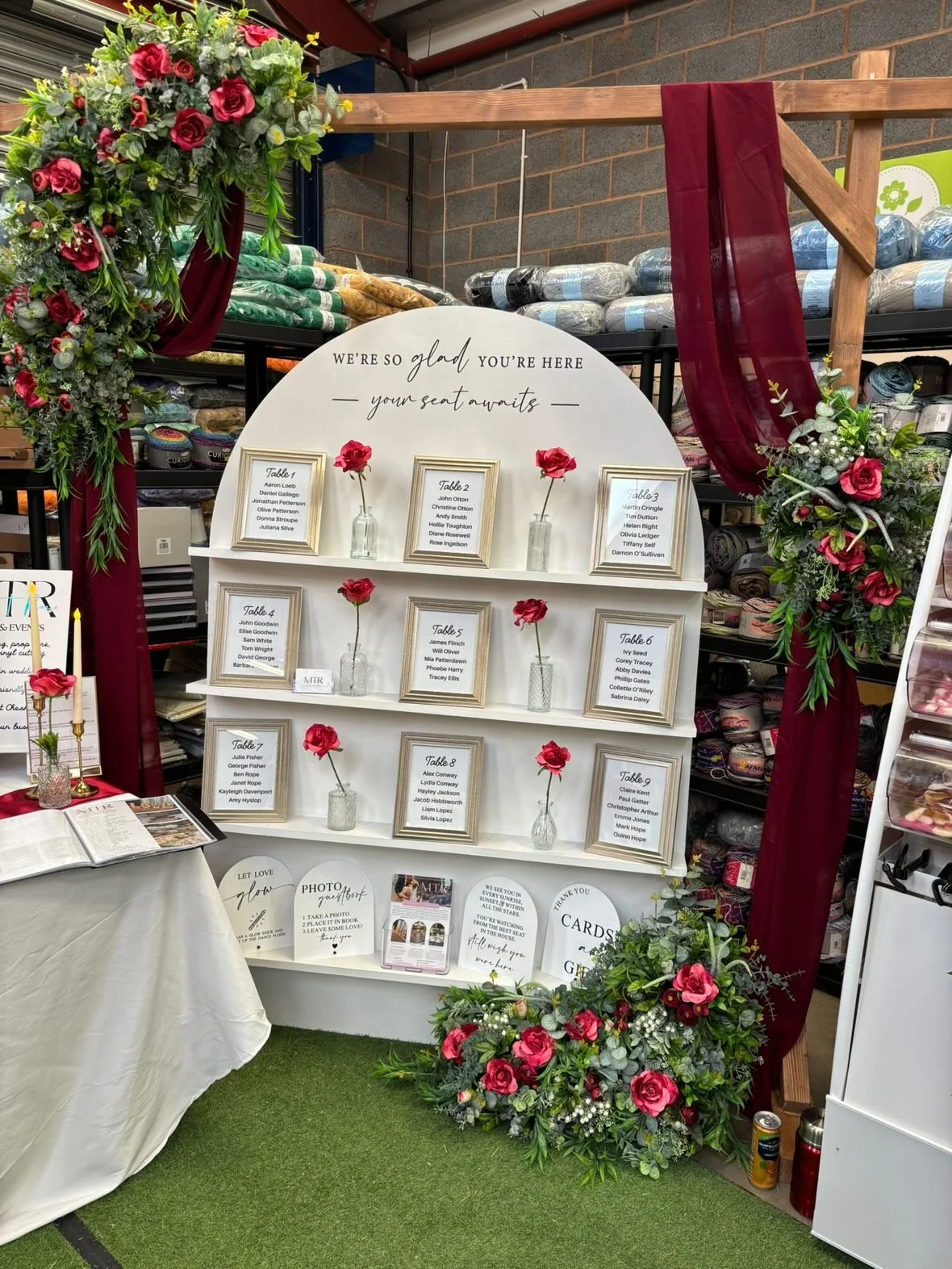 Decorative wedding seating chart display with white semi-circle board, framed name cards, single pink flowers in glass vases, surrounded by pink floral arrangements and burgundy drapes.