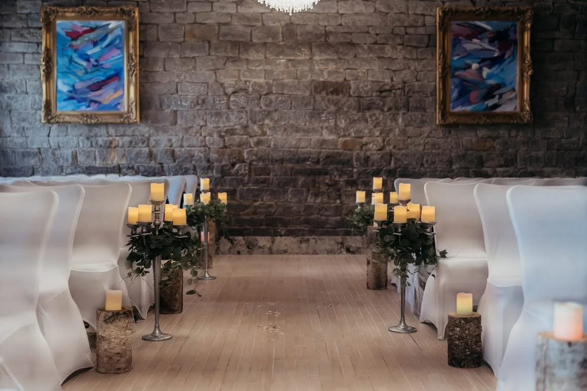 Wedding aisle decorated with candles and greenery, with white chairs covered in white fabric on both sides and abstract paintings hanging on a brick wall in the background.