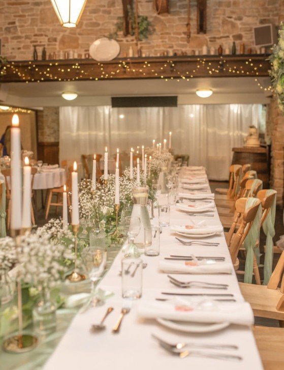 A long banquet table set for a formal event or celebration in a rustic-style venue decorated with candles, flowers, and fairy lights.