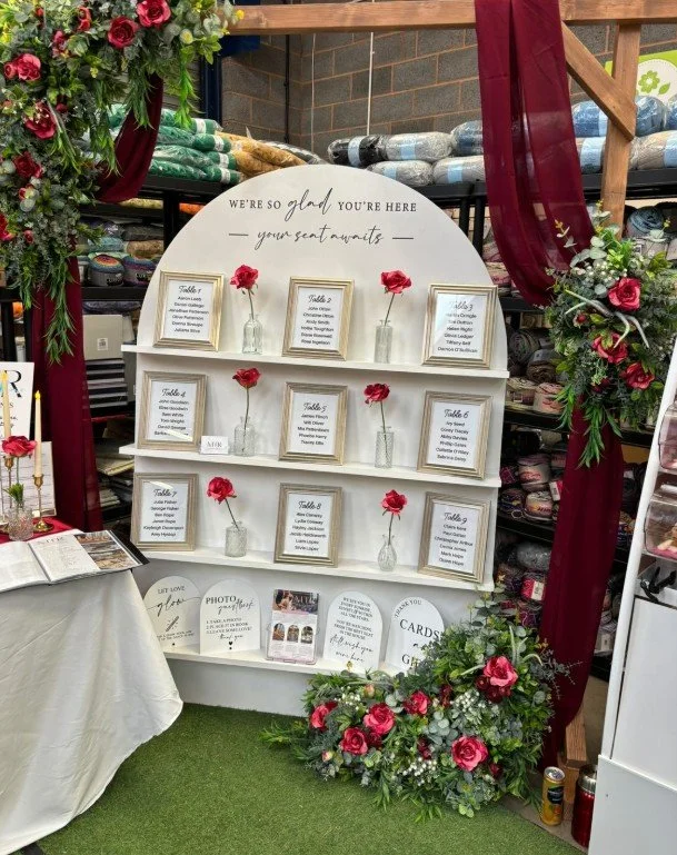 Wedding or event seating arrangement display with a white shelf decorated with pink roses in small glass vases, surrounded by floral arrangements and burgundy drapes.