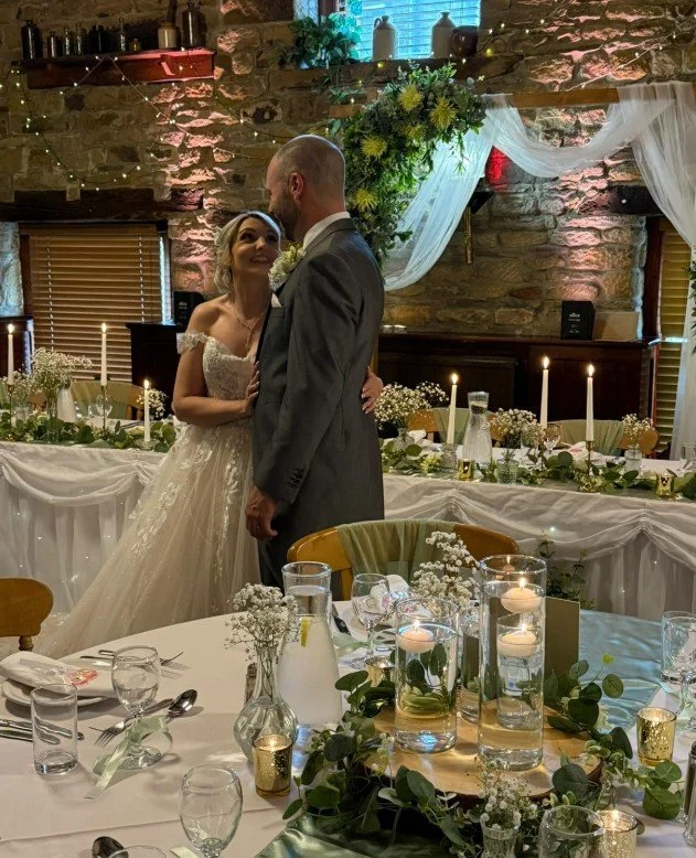 A bride and groom share a moment during their wedding reception, with the bride in a white gown and the groom in a gray suit, surrounded by decorated tables and candles.