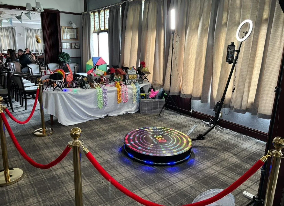 Photo of a decorated table with leis, flowers, and sign that says "Lovers" in a room with large windows and beige curtains, a ring light, a wireless phone tripod, and a dance pad on the floor with colorful LED lights, likely set up for a celebration 
