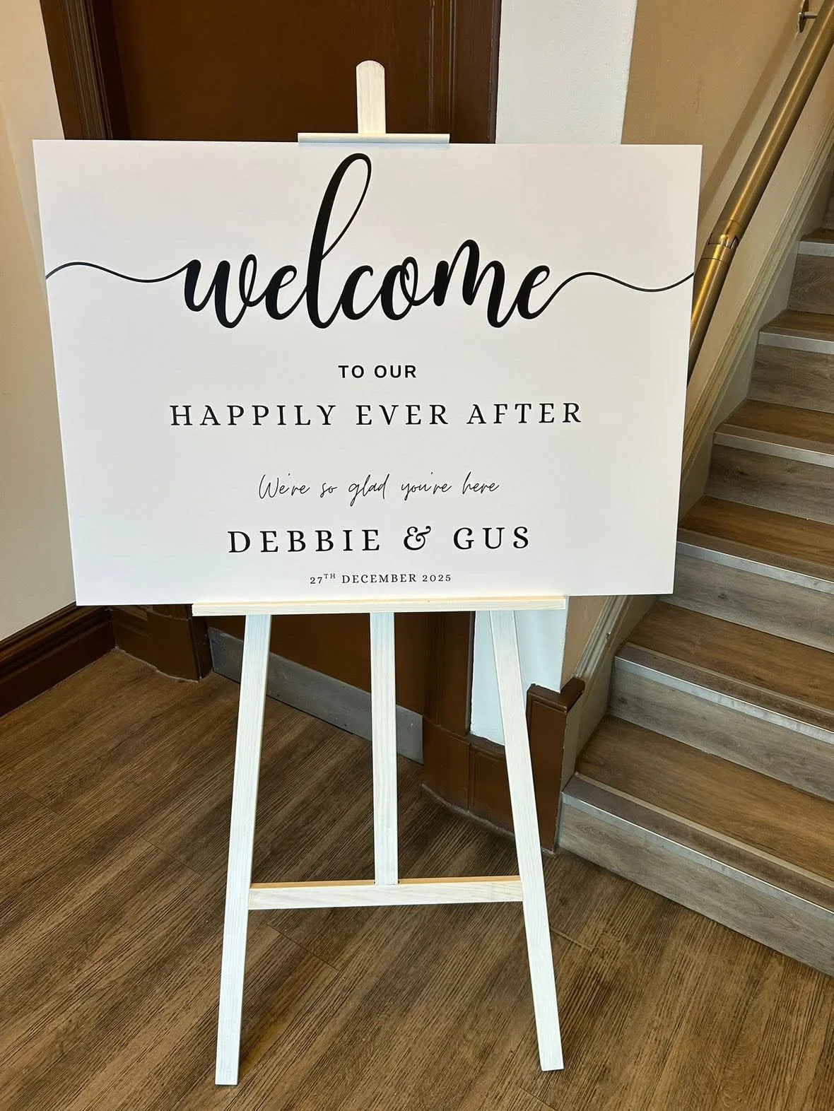 A white welcome sign on a wooden easel, decorated with black script and serif fonts, reading 'Welcome to our happily ever after. We're so glad you're here Debbie & Gus, 27th December 2025,' positioned near a staircase.