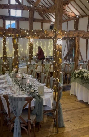 Decorated wedding reception area with long dining tables, white chairs, fairy lights, and rustic wooden beams in a barn-style venue.