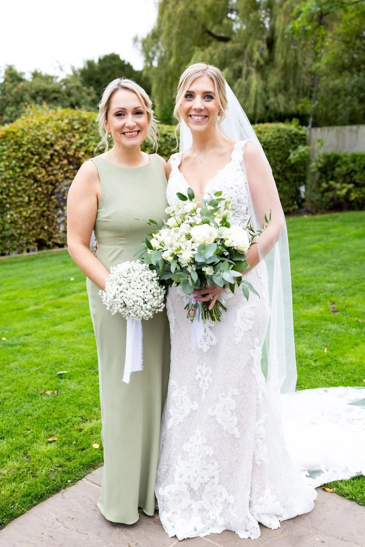 Two women dressed for a wedding, one in a white lace wedding gown with a veil, holding a bouquet, and the other in a light green bridesmaid dress, holding a small bouquet, standing outdoors on grass with trees and bushes in the background.