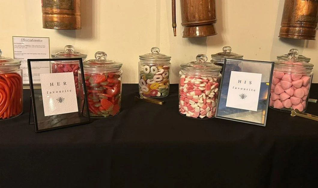 Jars filled with various candies, including gummy worms, gummy rings, marshmallows, and pink heart-shaped candies, on a black table, labeled 'HER favorite' and 'HIS favorite'.