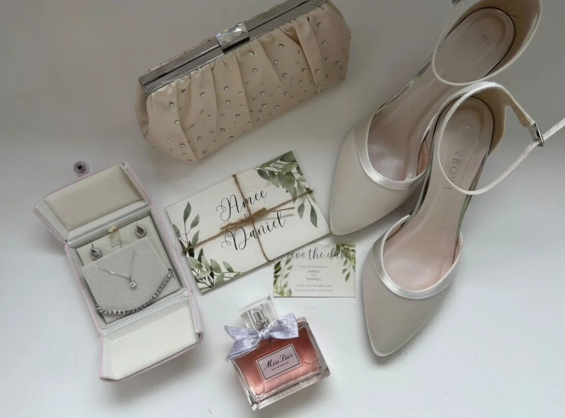 Wedding accessories including a pair of cream high-heeled shoes, a beige clutch purse with silver studs, a jewelry box with earrings and a necklace, a pink perfume bottle with a silver ribbon, and a wedding invitation with green leaves design.
