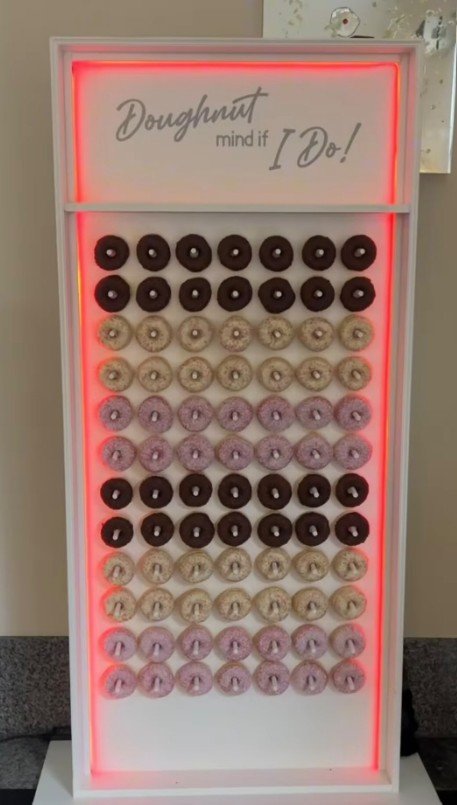 A donut display case with rows of chocolate and glazed donuts. The top row has chocolate donuts, followed by alternating glazed and pink frosted donuts with sprinkles. The display has a neon light border and a sign that reads, 'Doughnut mind if I do!