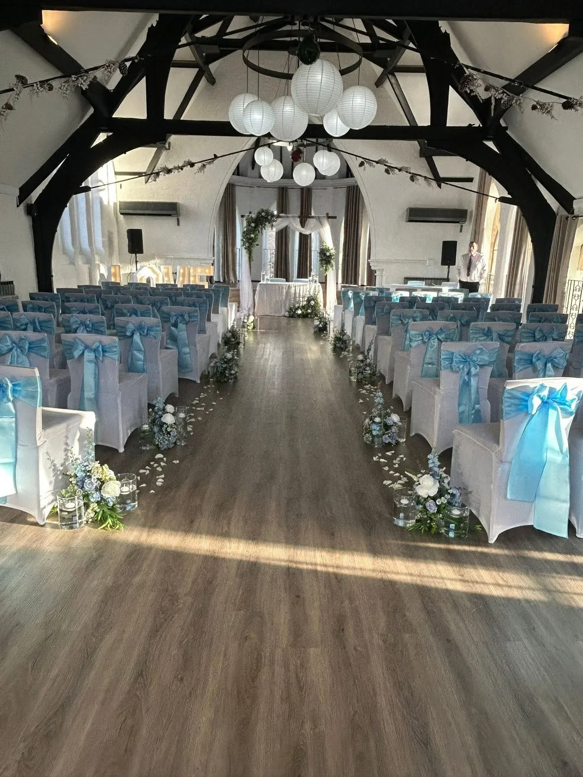 Wedding ceremony setup in a decorated hall with rows of white-covered chairs with blue ribbons, floral arrangements along the aisle, and a decorated altar at the front, under a vaulted ceiling with hanging white lanterns.