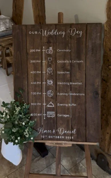 Wedding schedule sign on a wooden board with times and events for a wedding day, including ceremony, cocktails, speeches, breakfast, celebrations, buffet, carriages, and names with date.