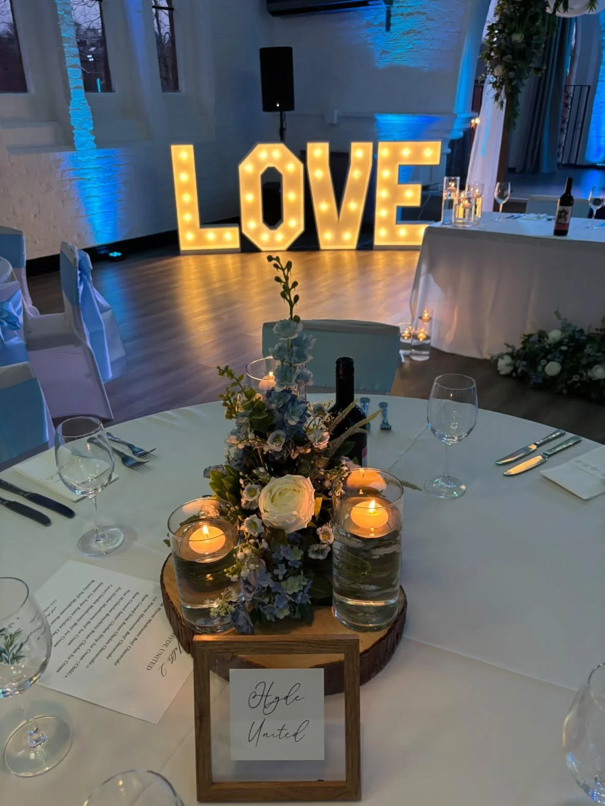 Decorated wedding reception table with a floral centerpiece, candles, wine bottles, and glasses, with large illuminated love sign in the background.