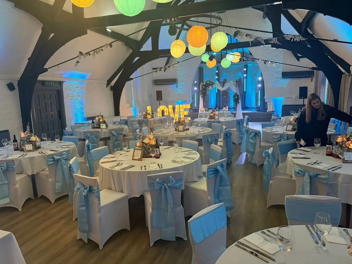 Decorated banquet hall with round tables covered in white tablecloths, each adorned with floral centerpieces, glasses, and table settings. Chairs are wrapped in white covers with blue bows. Colorful paper lanterns hang from the ceiling, and large ill