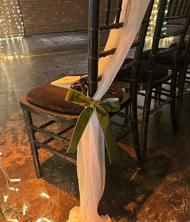 Black Chiavari chairs decorated with cream-colored tulle and green satin ribbons tied in bows for a wedding or special event, with string lights on the floor.