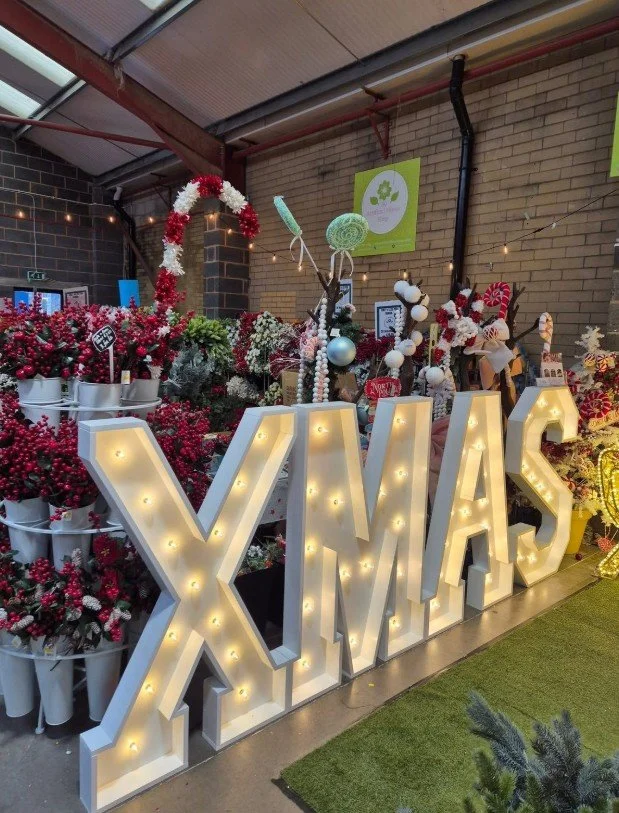 Decorative Christmas display with large illuminated sign spelling 'XMAS' surrounded by Christmas trees, ornaments, and festive decorations in an indoor setting.