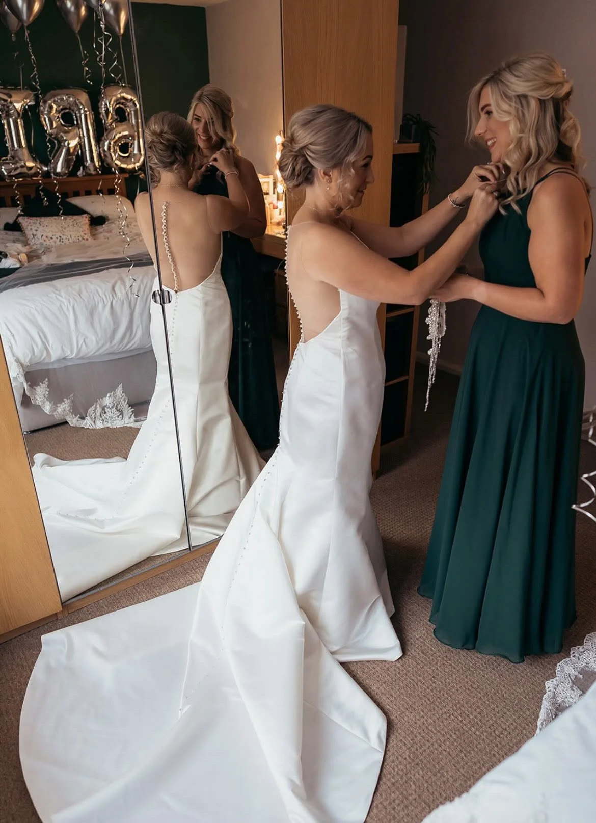 A bride in a white wedding gown with a long train adjusts a bridesmaid's green dress in front of a mirror, with balloons and party decorations in the background.