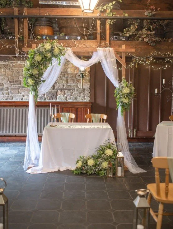 Wedding or special event ceremony setup with a decorated arch, white drapes, flowers, candles, and lanterns in a rustic indoor venue.
