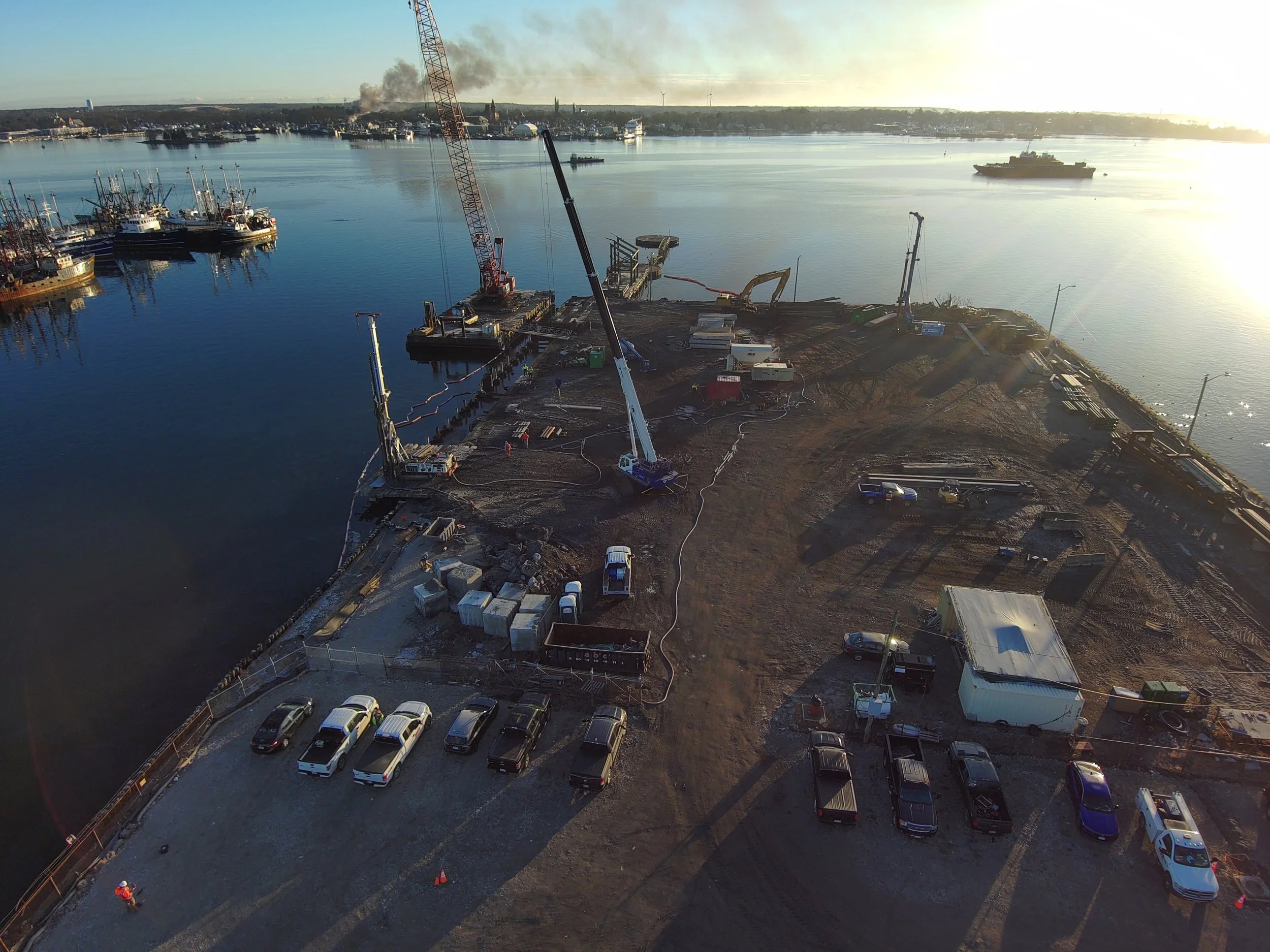 Construction site on a waterfront with cranes, construction vehicles, and parked cars, adjacent to a harbor with docked boats and ships, and smoke rising in the background at sunset.