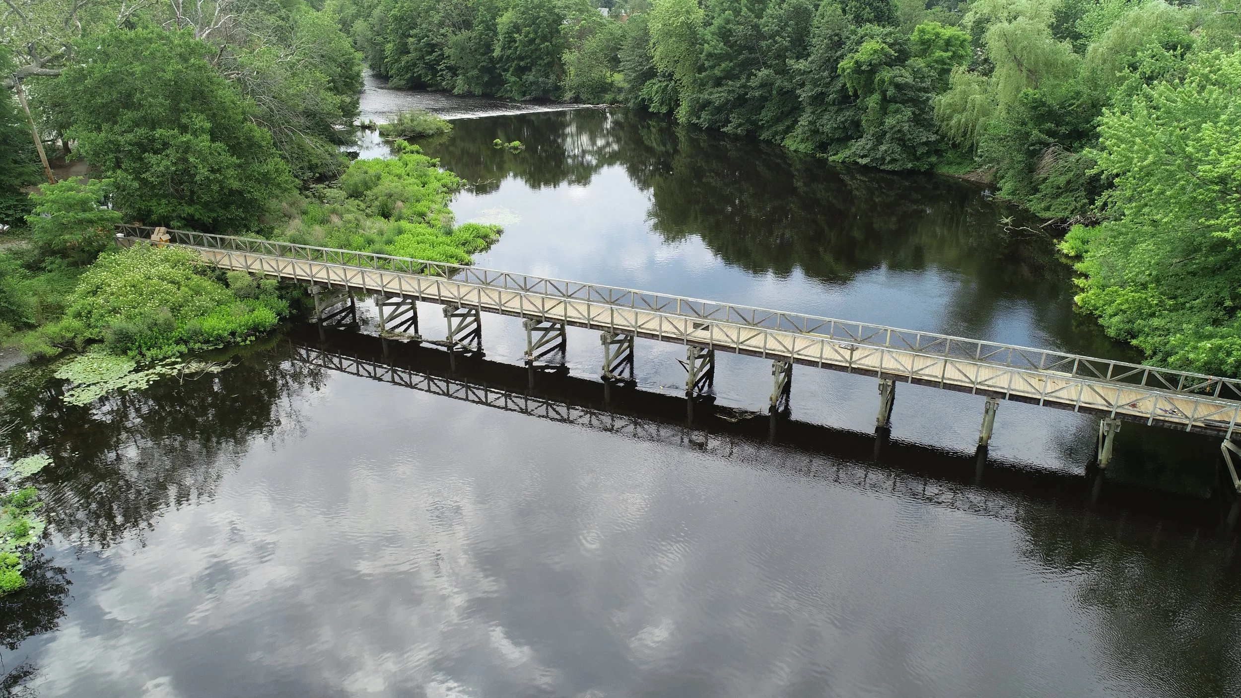 An aerial view of a wooden bridge crossing a calm river surrounded by lush green trees.