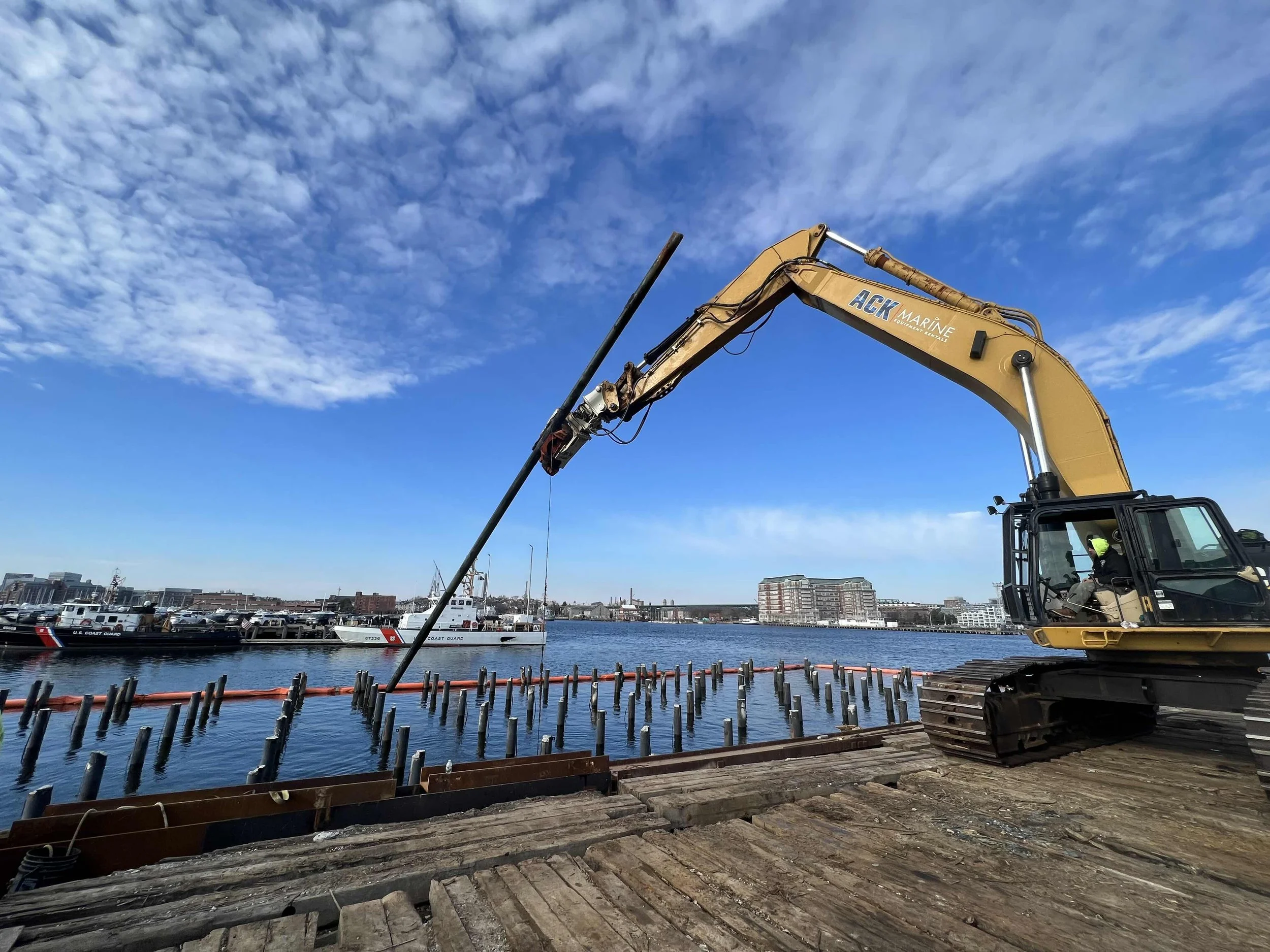 Construction worker operating a yellow excavator at a dock by a harbor with boats, under a partly cloudy sky.