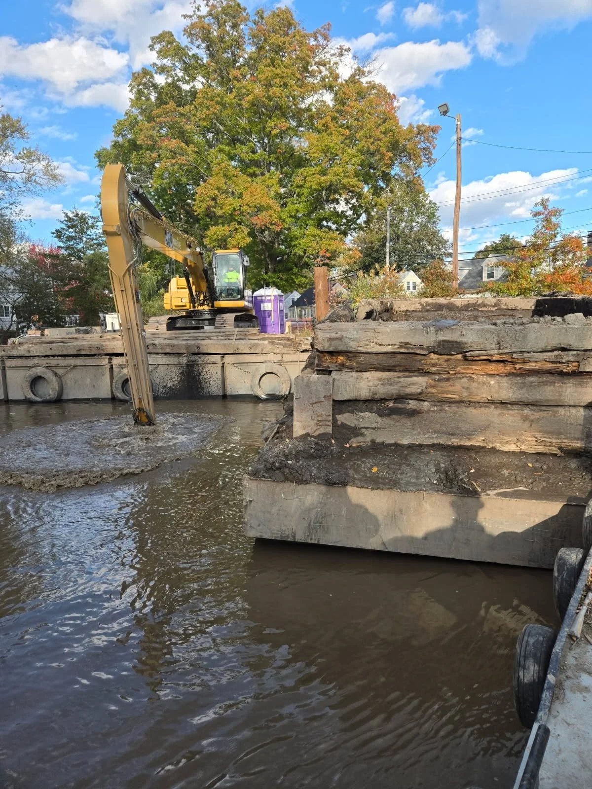 Construction work on a waterway using a mini excavator, with large wood and concrete beams, trees with fall foliage, houses in the distance, and a bright, partly cloudy sky.