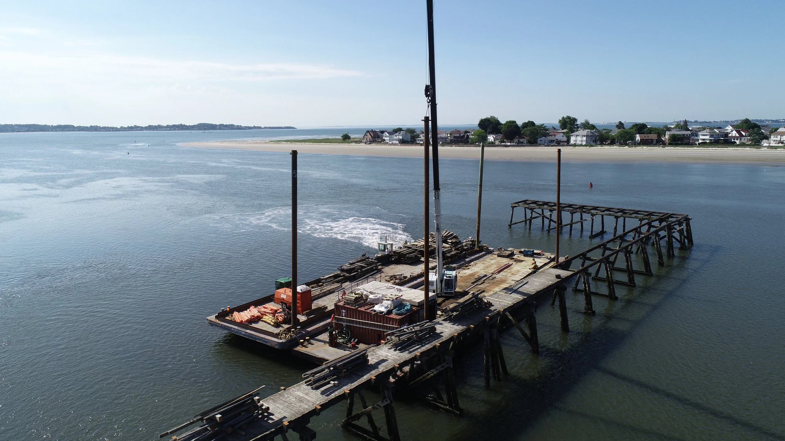 Construction site on a partially demolished wooden pier in a body of water, with construction equipment and materials on the pier, and a residential shoreline in the background.